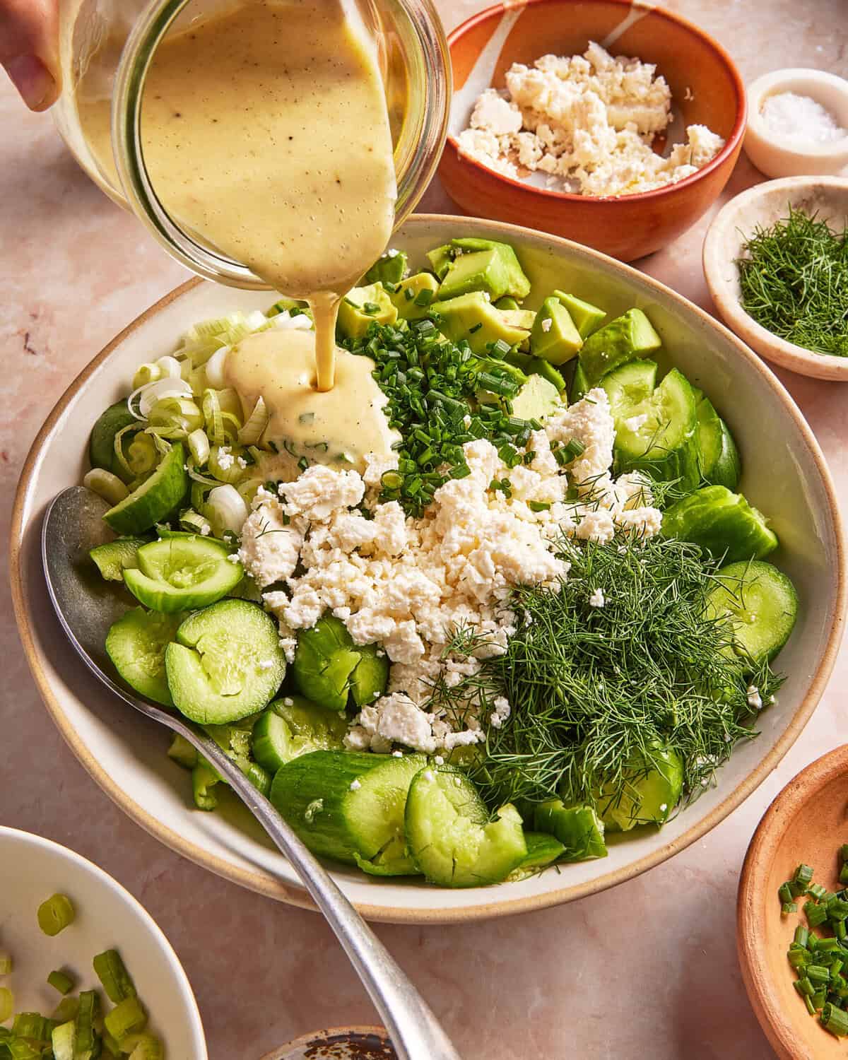 A hand pours creamy dressing onto a salad with sliced cucumbers, crumbled feta, fresh dill, chopped chives, and green onions in a large bowl. Small bowls of feta, dill, and salt surround the salad.