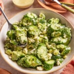 A bowl of cucumber salad with crumbled feta cheese and fresh herbs, served with a spoon. A jar of creamy dressing, utensils, and small dishes with herbs are nearby on a light-colored surface.