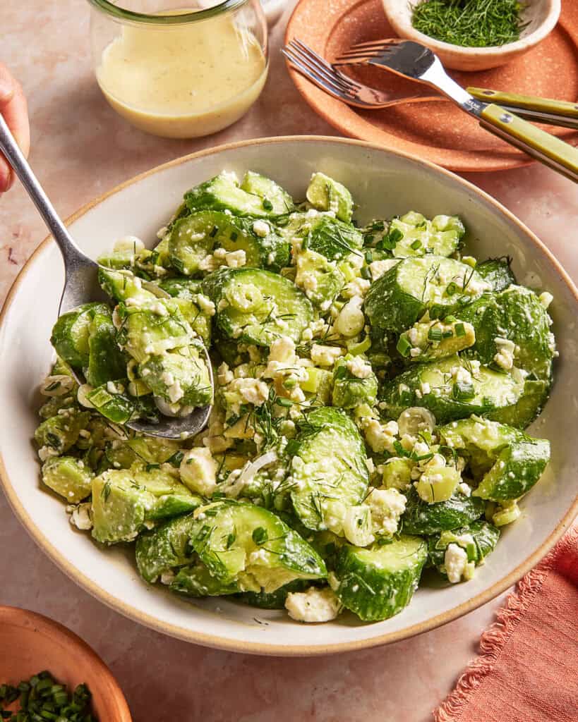 A bowl of cucumber salad with crumbled feta cheese and fresh herbs, served with a spoon. A jar of creamy dressing, utensils, and small dishes with herbs are nearby on a light-colored surface.
