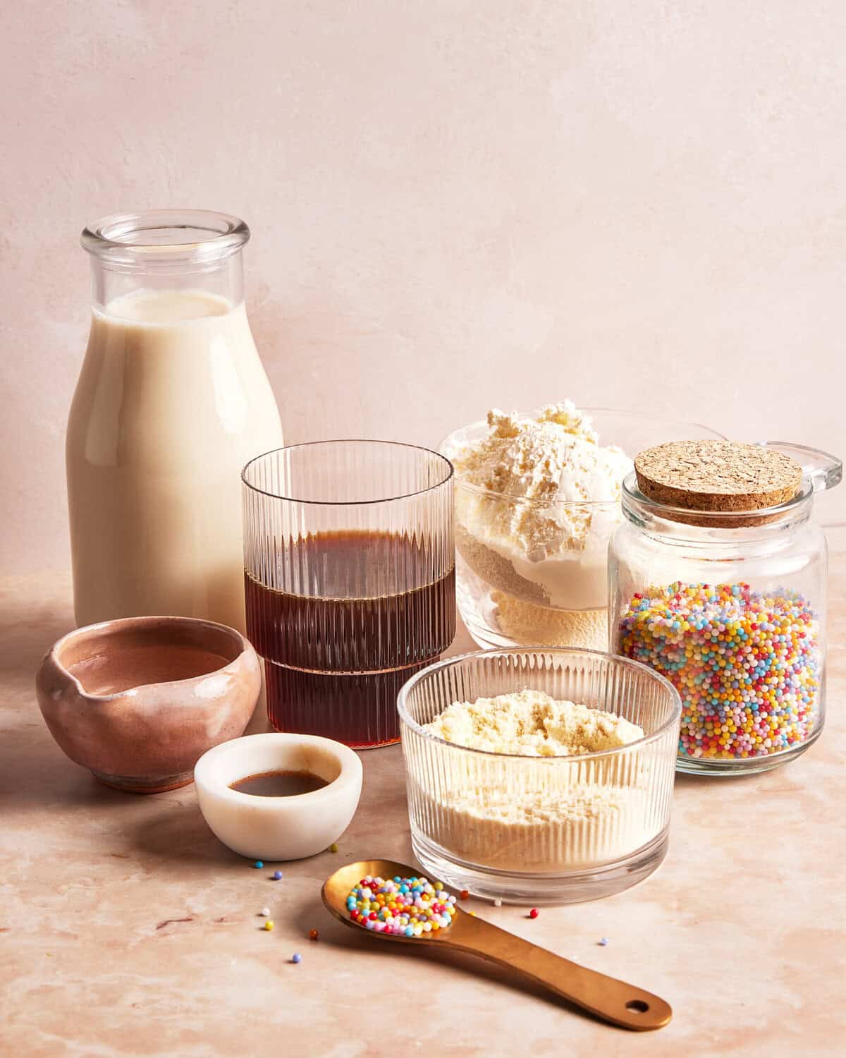 A variety of baking ingredients on a light counter, including a glass bottle of milk, a glass of dark liquid, bowls of flour and sugar, a jar of sprinkles, a wooden spoon with sprinkles, and small bowls with liquid and powder.