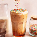 A glass of iced coffee topped with colorful round sprinkles, being poured from a spoon. A jar of sprinkles, a glass of milk, and a brown glass are nearby on a light background.