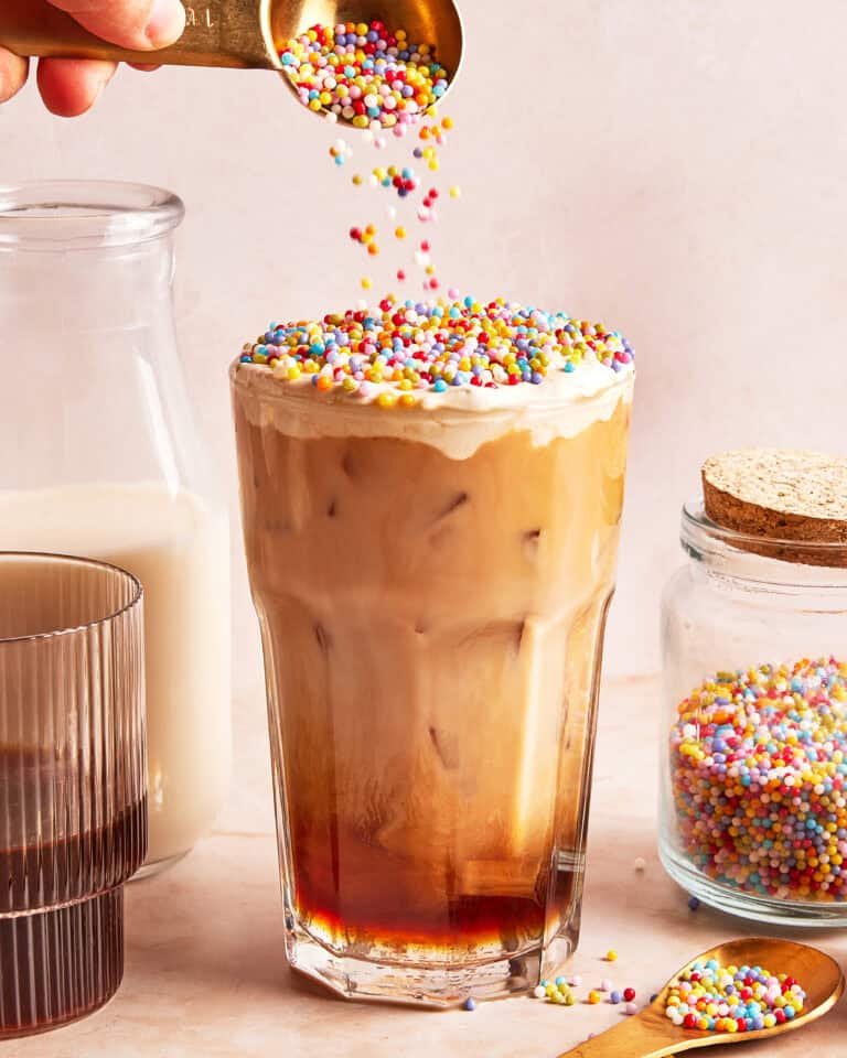 A glass of iced coffee topped with colorful round sprinkles, being poured from a spoon. A jar of sprinkles, a glass of milk, and a brown glass are nearby on a light background.
