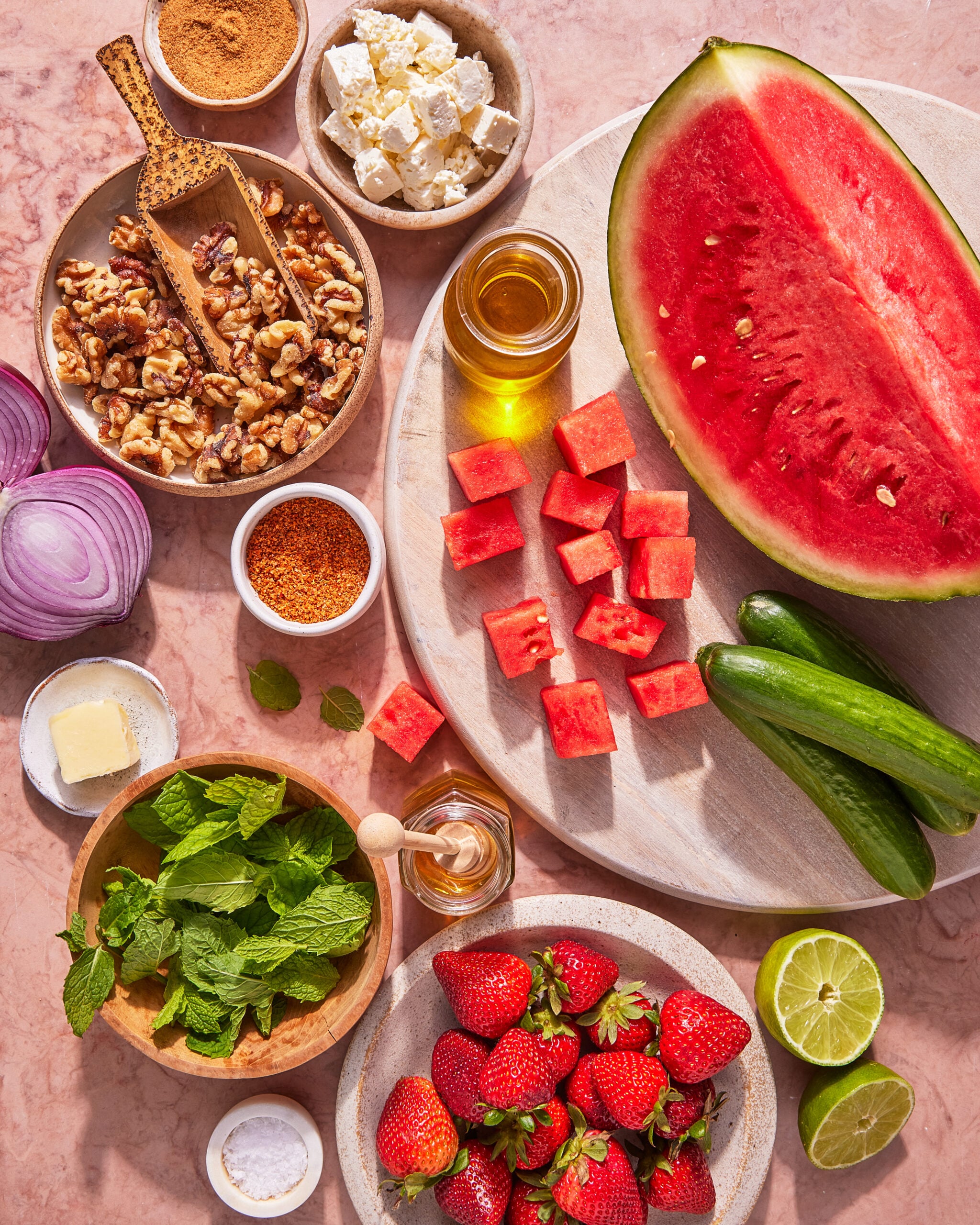 Watermelon cubes, cucumber, strawberries, lime halves, walnuts, feta cheese, red onion slices, fresh mint, honey, butter, oil, a seasoning blend, and salt arranged on a pink marble surface.