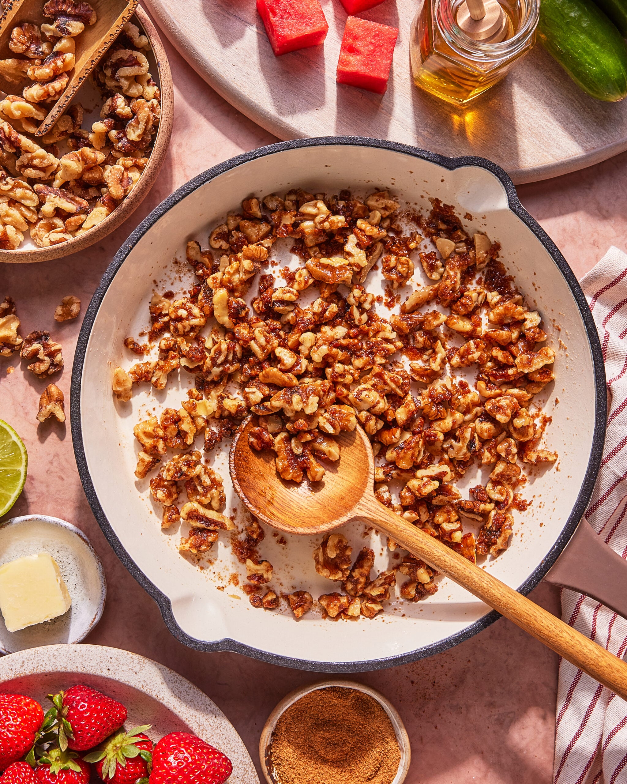 A skillet with toasted walnuts stirred by a wooden spoon, surrounded by strawberries, watermelon cubes, sugar, a lime, butter, walnuts, a jalapeño, and a bottle of olive oil on a pink surface.