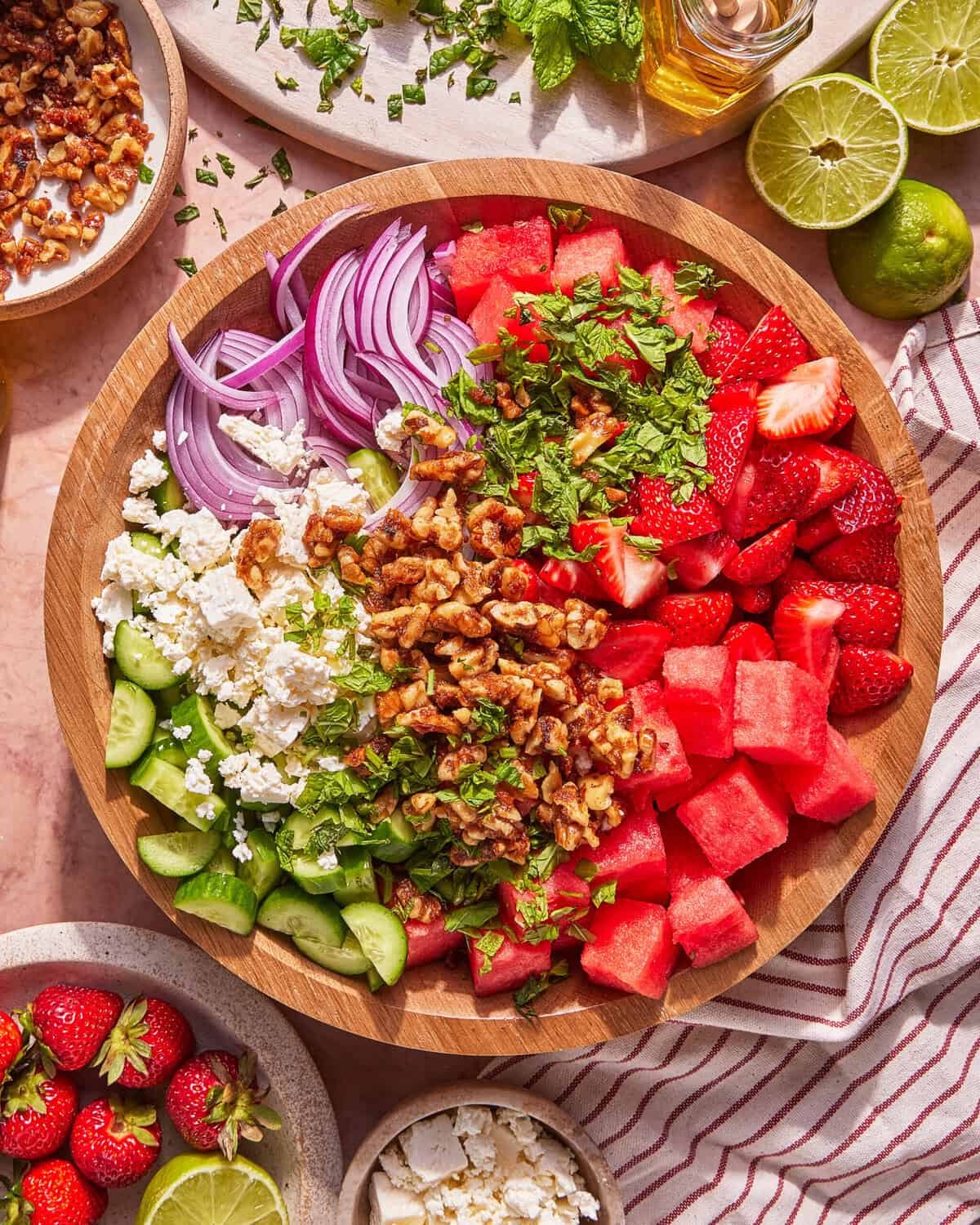 A wooden bowl filled with watermelon cubes, sliced strawberries, cucumber, feta cheese, red onion, chopped mint, and walnuts, arranged in separate sections. Plates with more ingredients and a striped napkin surround the bowl.