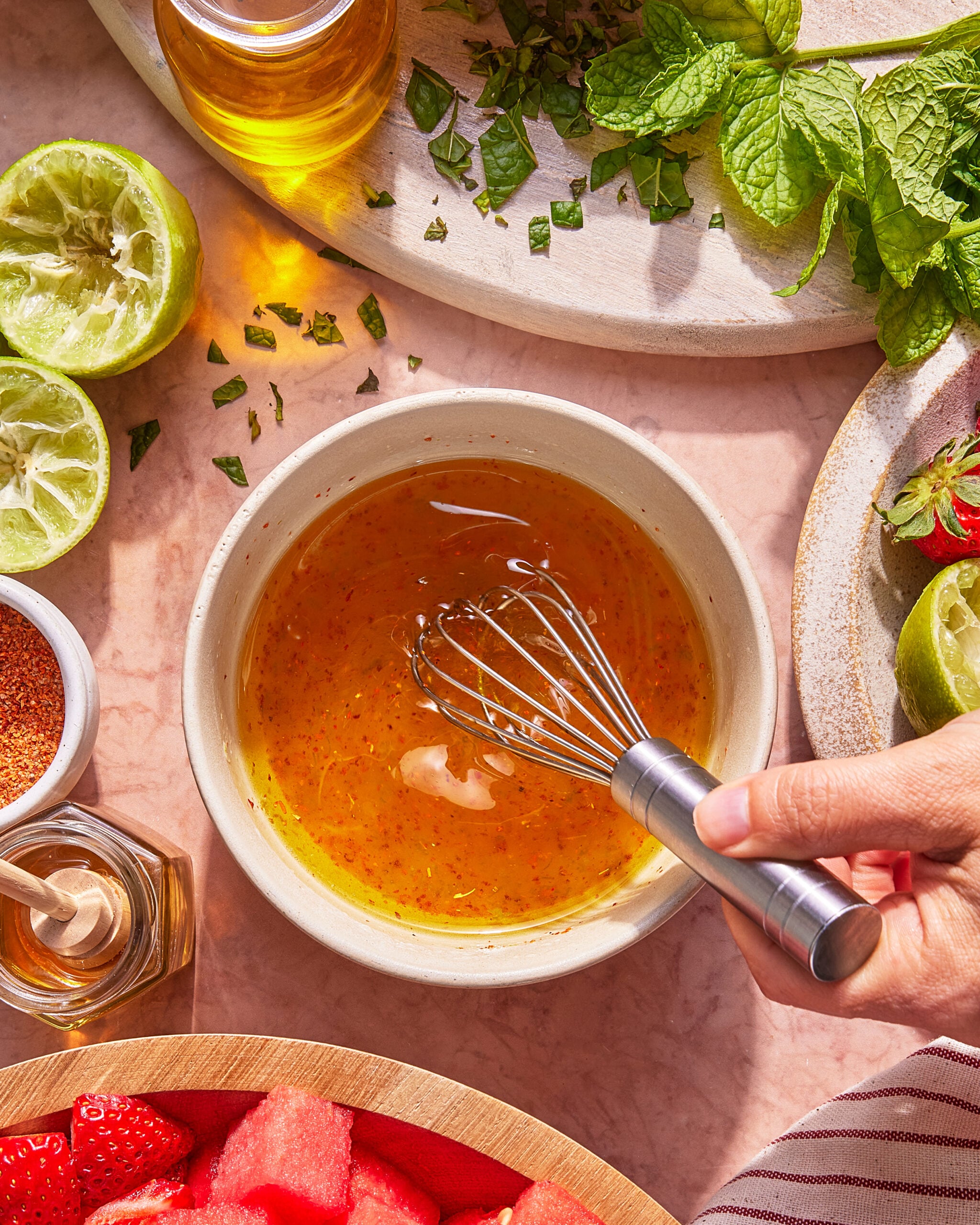 A hand whisking a golden liquid in a bowl surrounded by halved limes, fresh mint, a jar of honey, sliced strawberries, and a bowl of watermelon on a light pink surface.