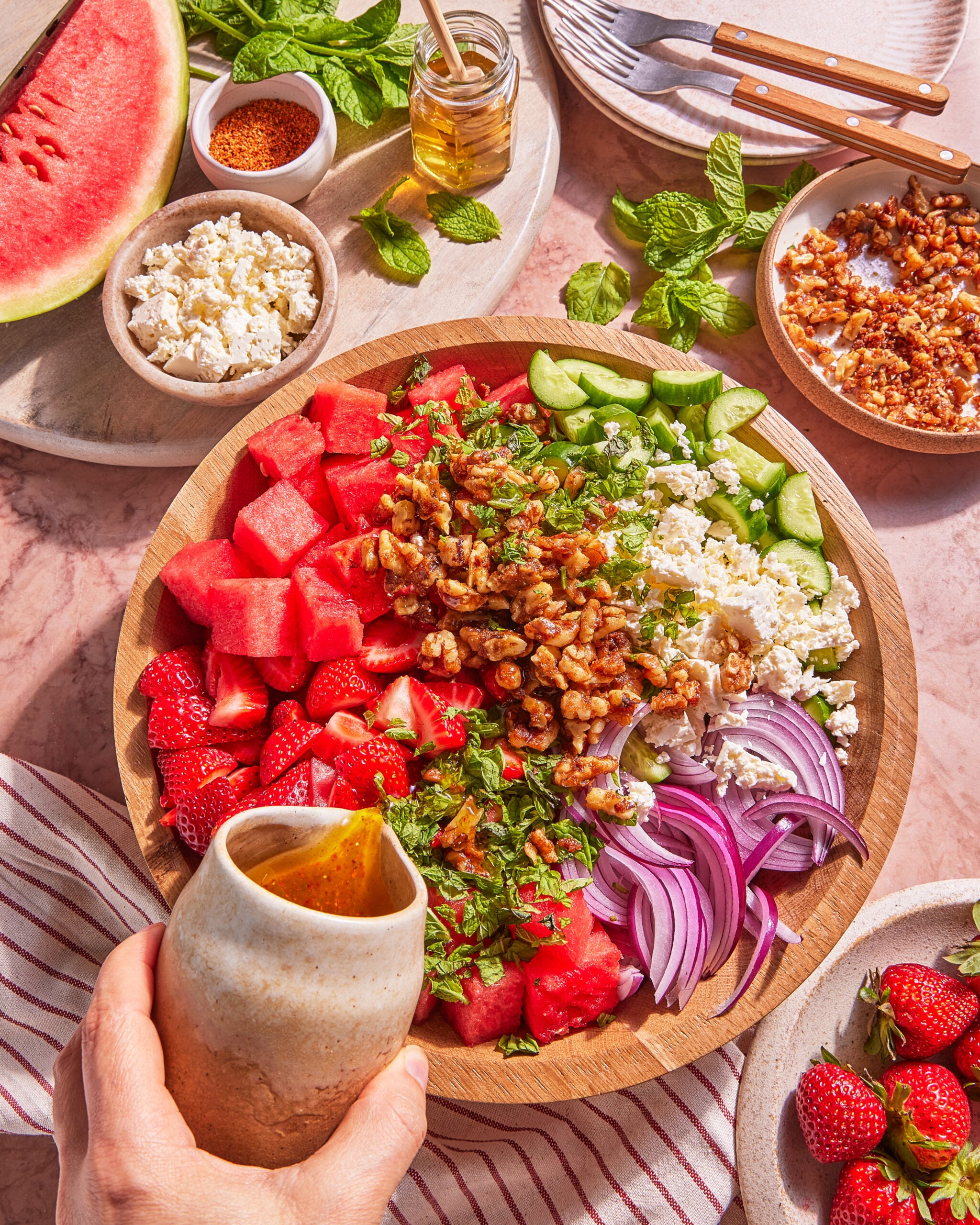 A hand pours dressing onto a vibrant salad with watermelon, strawberries, red onion, cucumber, feta, walnuts, and mint in a wooden bowl. Nearby are bowls of feta, walnuts, chili powder, and a slice of watermelon.
