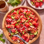 A wooden bowl filled with a colorful salad of watermelon, strawberries, cucumber, red onion, feta cheese, and nuts, with a serving spoon. A plate with salad, mint leaves, and a watermelon slice are nearby.