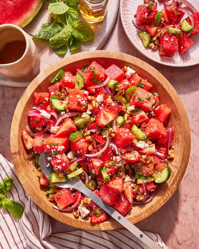 A wooden bowl filled with a colorful salad of watermelon, strawberries, cucumber, red onion, feta cheese, and nuts, with a serving spoon. A plate with salad, mint leaves, and a watermelon slice are nearby.