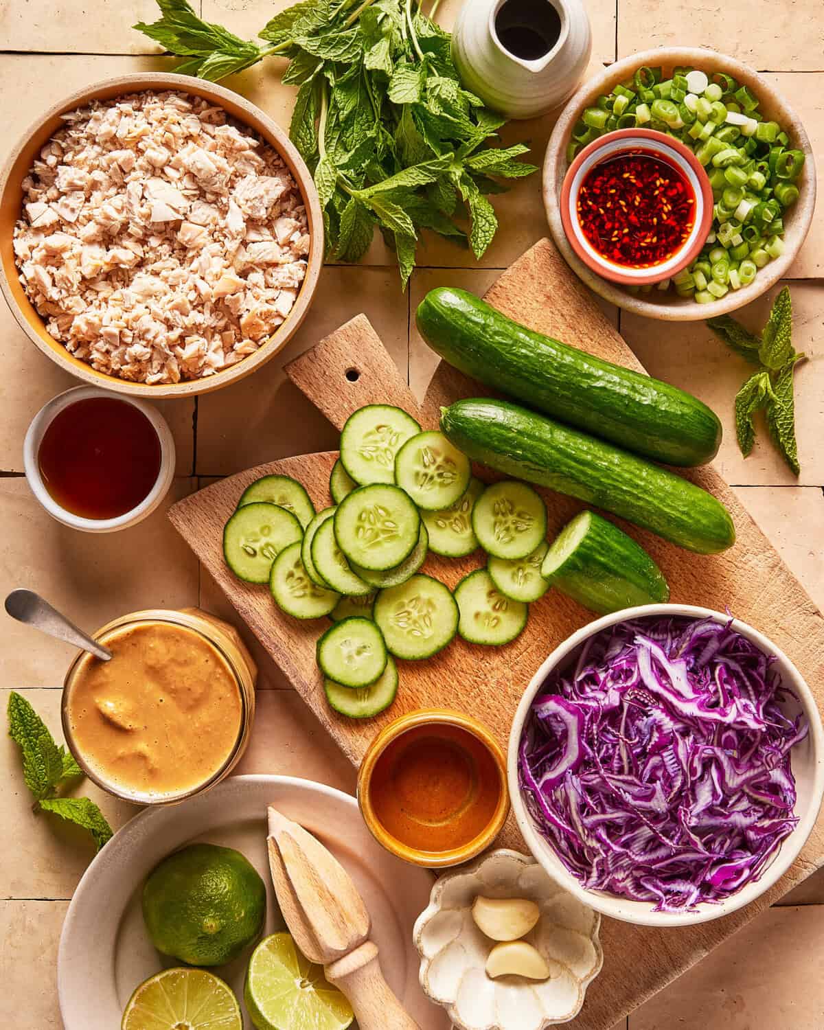 Top-down view of various fresh ingredients, including cucumbers, shredded cabbage, chopped chicken, sliced lime, garlic, sauces, green onions, mint leaves, and peanut butter, arranged neatly on a kitchen surface.