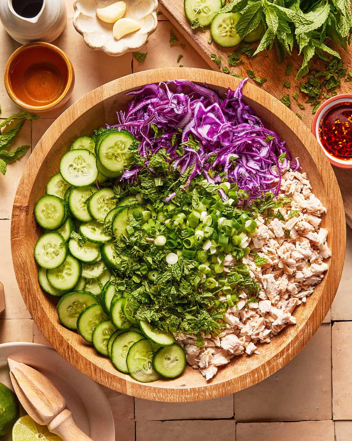 A wooden bowl filled with sliced cucumbers, shredded purple cabbage, chopped green onions, herbs, and cooked chicken sits on a tiled surface surrounded by ingredients.