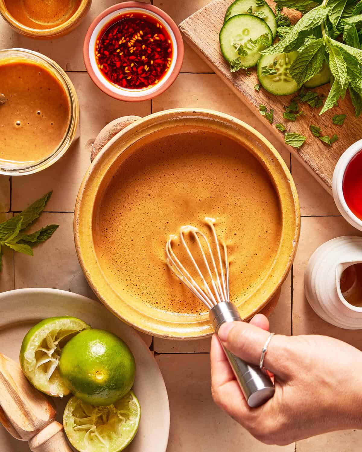 A hand whisks a creamy brown sauce in a bowl, surrounded by limes, herbs, sliced cucumber, chili oil, and various condiments on a tiled surface.
