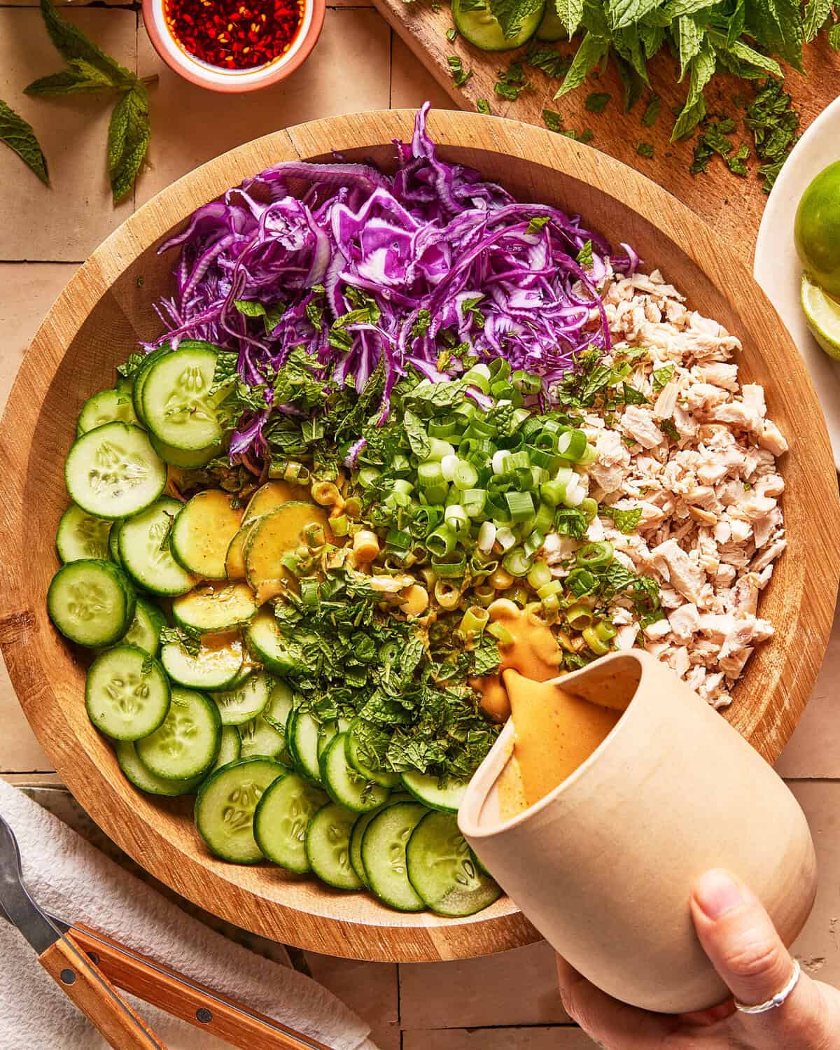 A hand pours dressing over a salad with sliced cucumbers, shredded purple cabbage, chopped chicken, green onions, and herbs arranged in a wooden bowl.