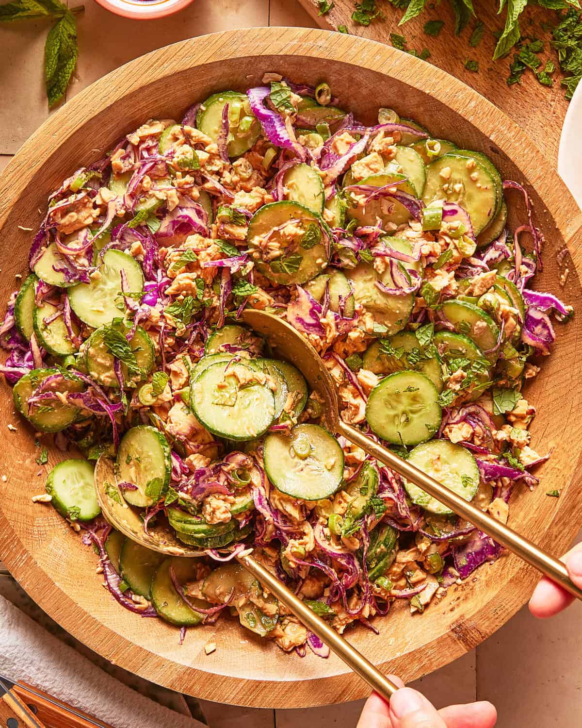 A wooden bowl filled with cucumber salad, red cabbage, fresh herbs, and chopped nuts, with two serving spoons mixing the ingredients.
