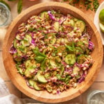 A wooden bowl filled with salad made of cucumber, red cabbage, peanuts, herbs, and mint leaves. The bowl is on a tiled surface with lime slices, mint, dipping sauce, and glasses of water nearby.