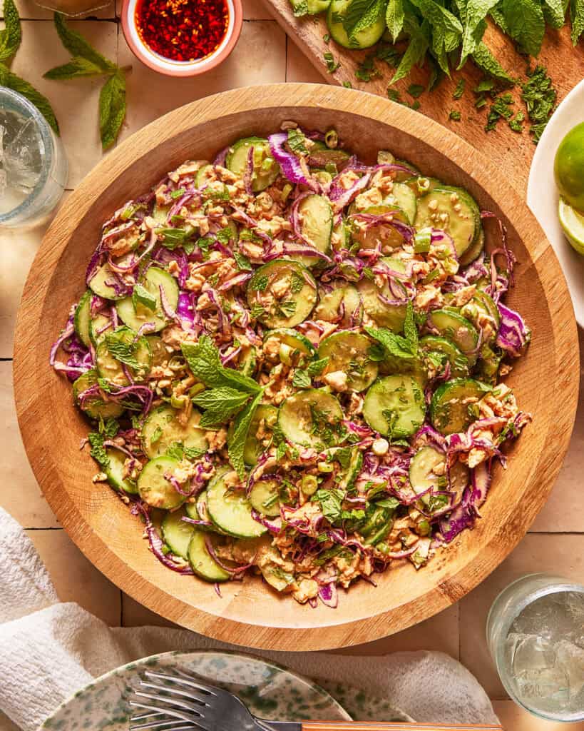 A wooden bowl filled with salad made of cucumber, red cabbage, peanuts, herbs, and mint leaves. The bowl is on a tiled surface with lime slices, mint, dipping sauce, and glasses of water nearby.