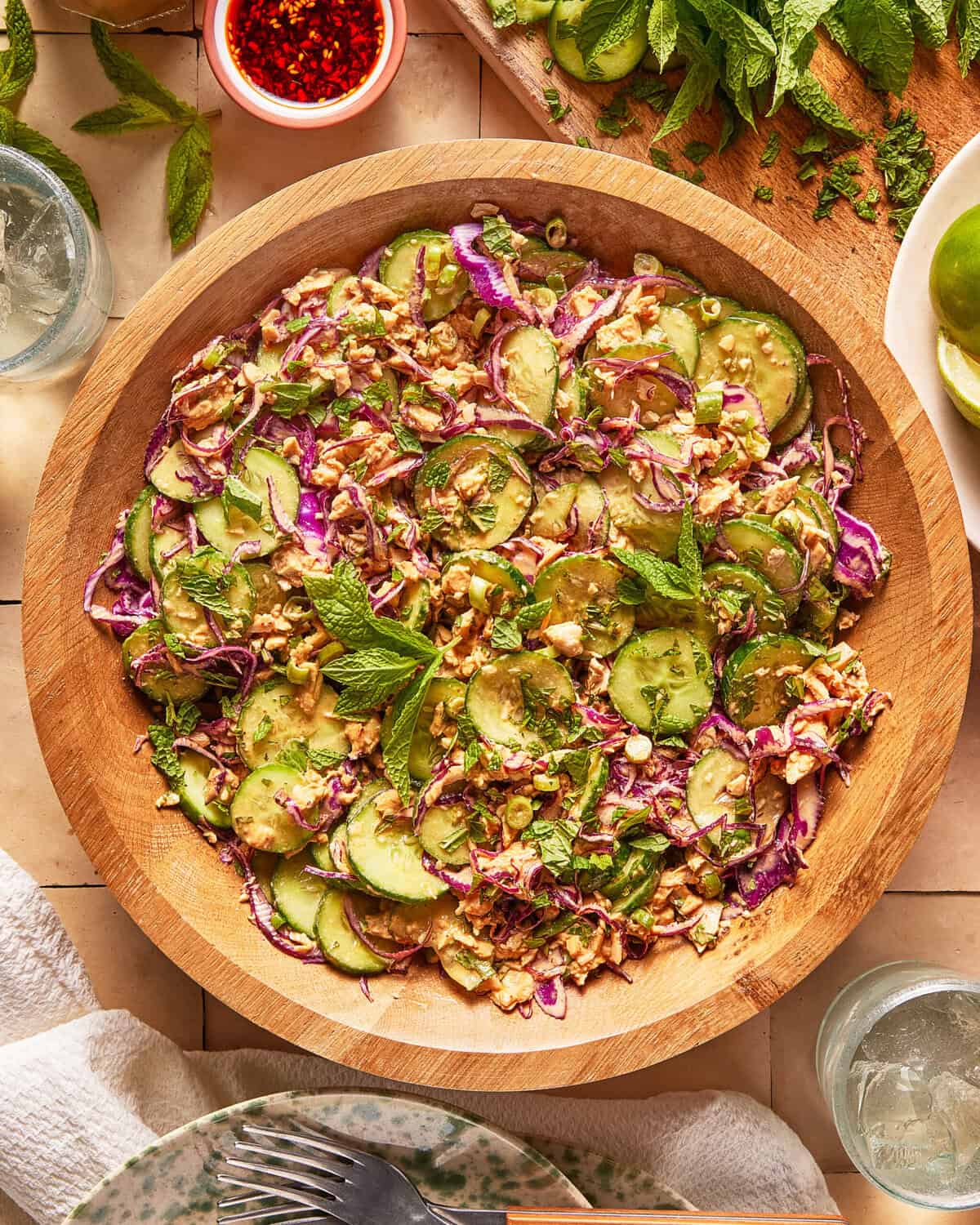 A wooden bowl filled with salad made of cucumber, red cabbage, peanuts, herbs, and mint leaves. The bowl is on a tiled surface with lime slices, mint, dipping sauce, and glasses of water nearby.