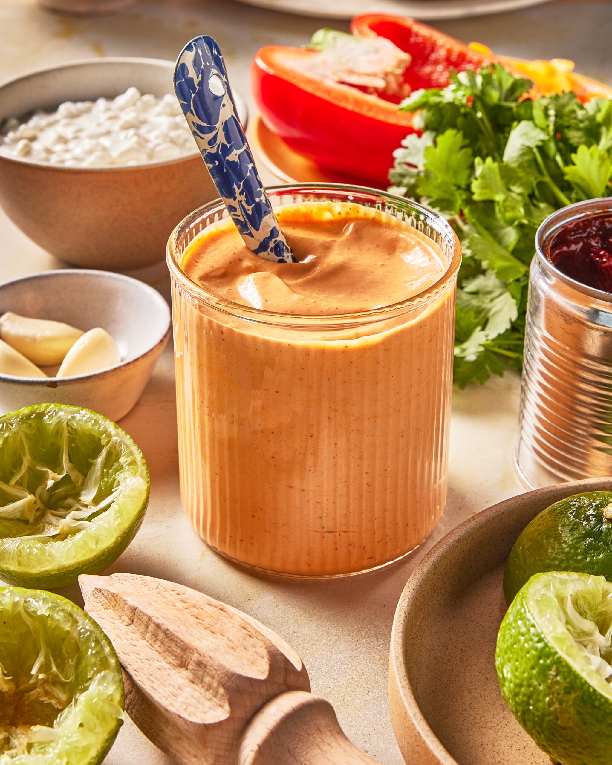 A glass of creamy orange sauce with a spoon, surrounded by halved limes, garlic, cottage cheese, cilantro, a red bell pepper, and a can of sauce on a kitchen counter.