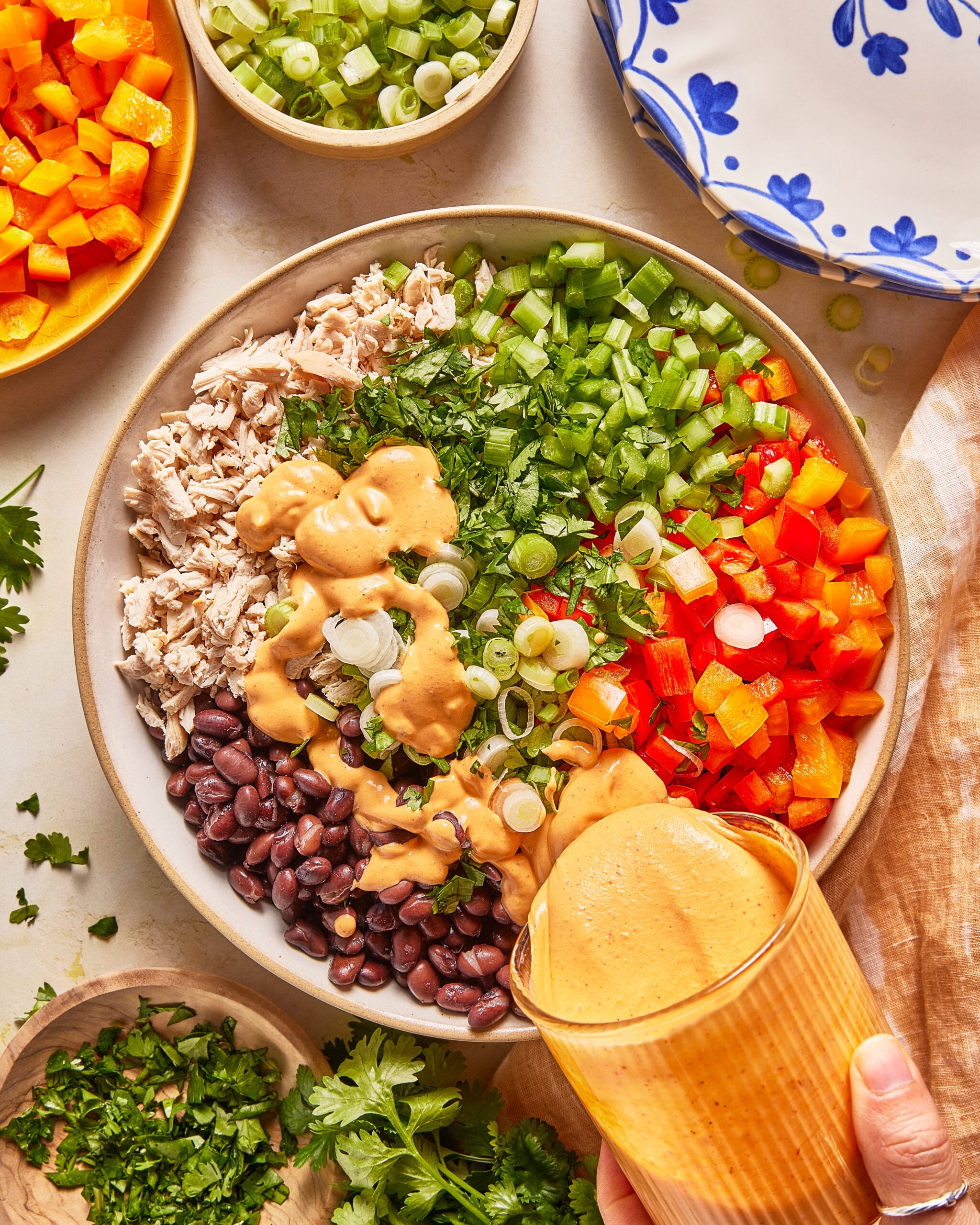 A bowl with shredded chicken, black beans, chopped celery, red and orange bell peppers, green onions, and cilantro, topped with creamy dressing. A hand pours more dressing from a glass. Plates and herbs surround the bowl.