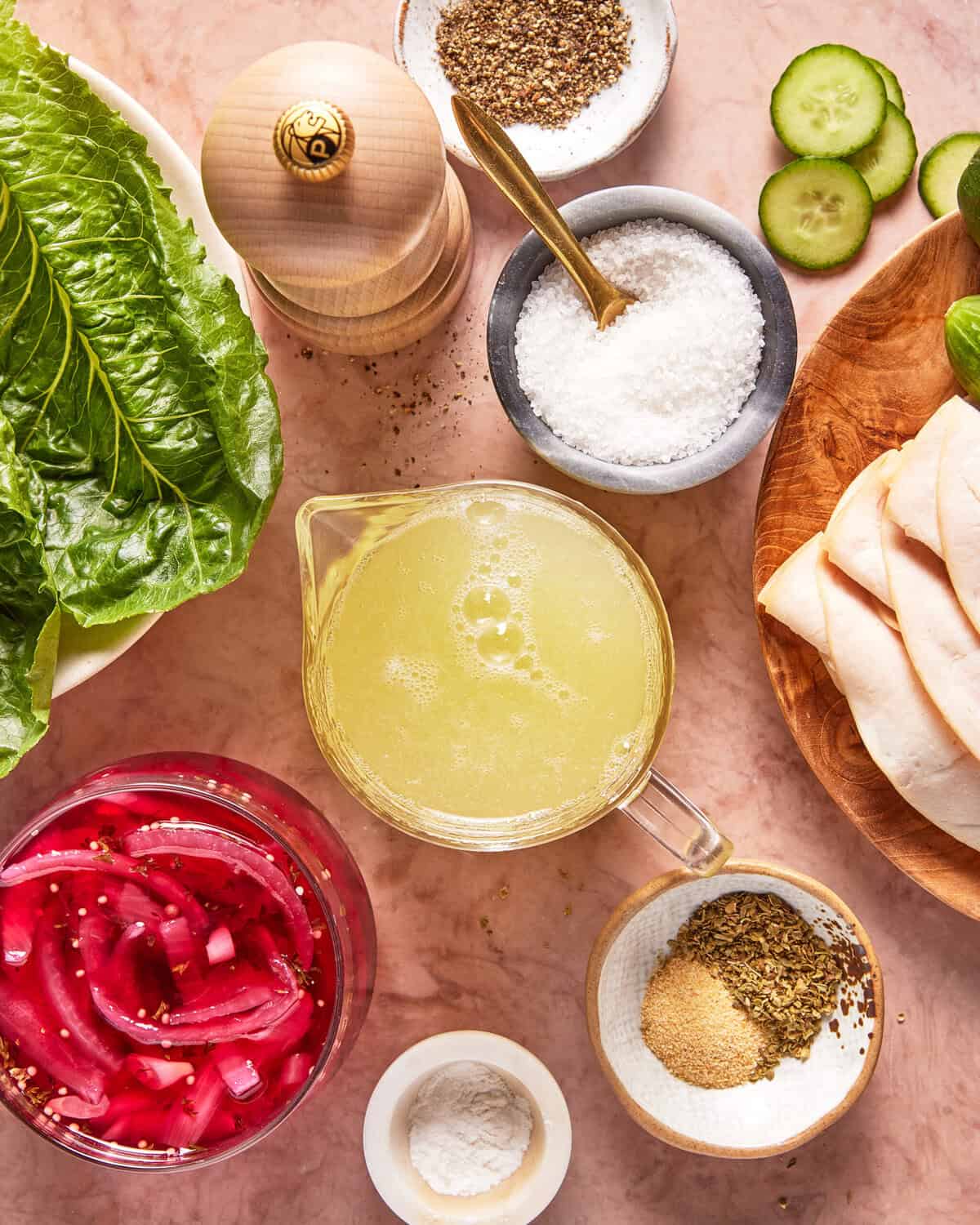 Ingredients on a pink surface including romaine leaves, pickled onions, salt, pepper, oregano, cucumber slices, flatbread, and a measuring cup of liquid.