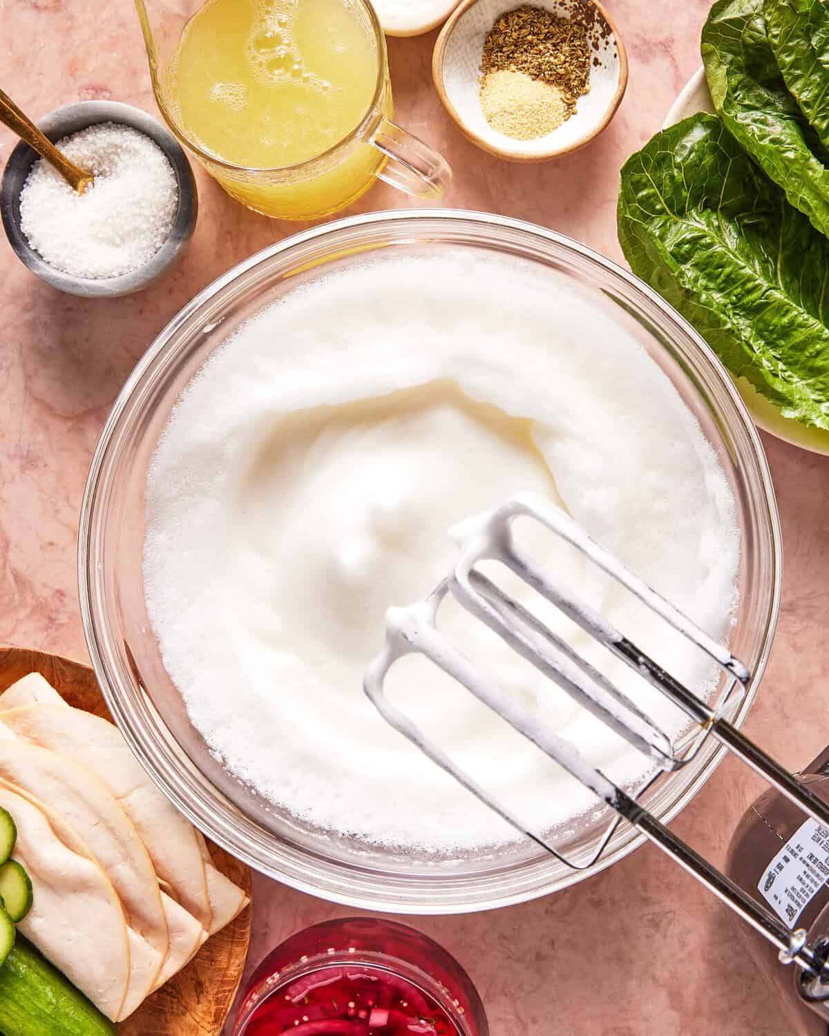 A bowl of whipped egg whites with a hand mixer surrounded by lettuce, sliced chicken, cucumber, pickled onions, broth, salt, and seasonings on a pink countertop.