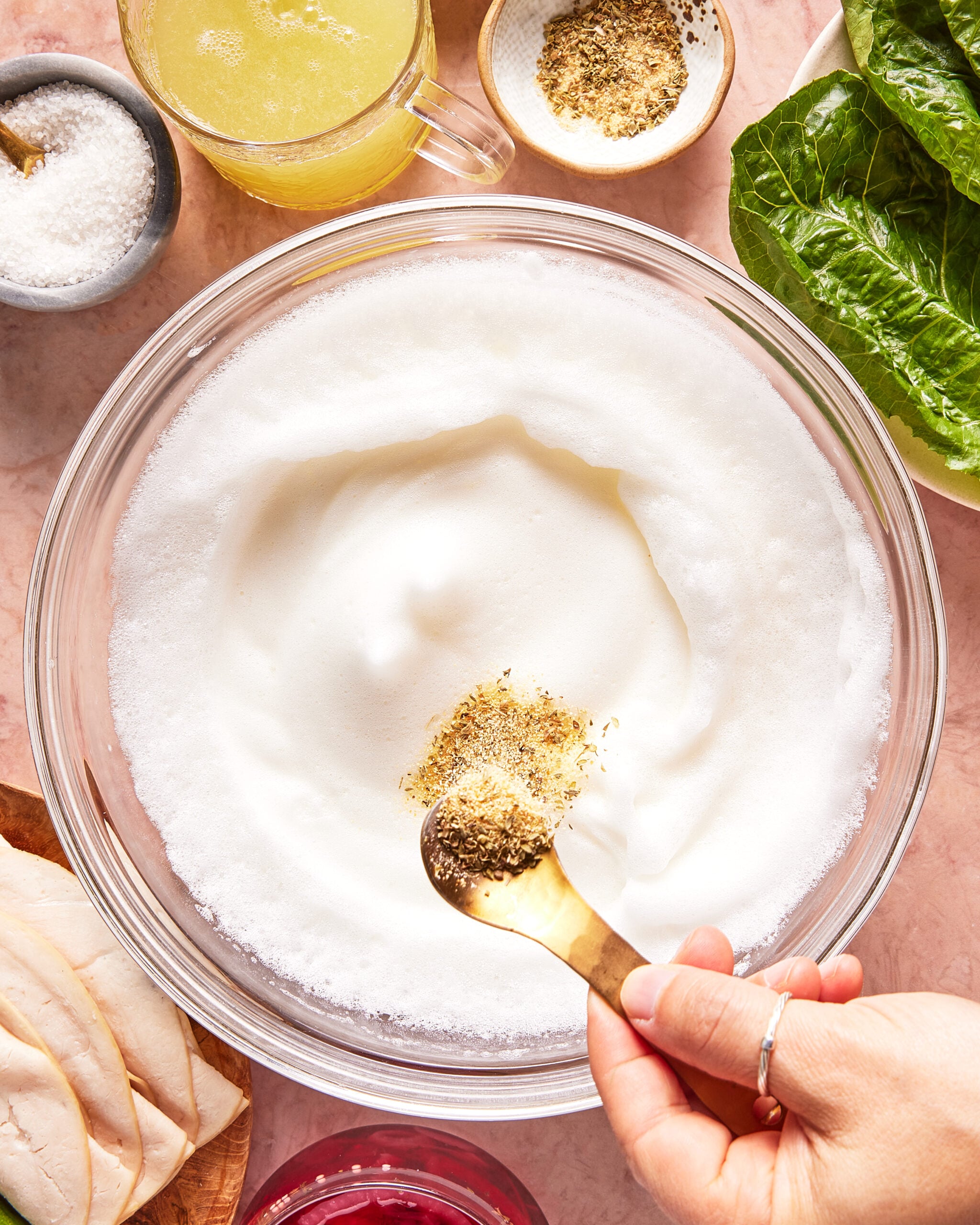 A hand adds a spoonful of dried herbs to a bowl of whipped egg whites, surrounded by salt, broth, lettuce, chicken slices, and a pink drink on a light surface.