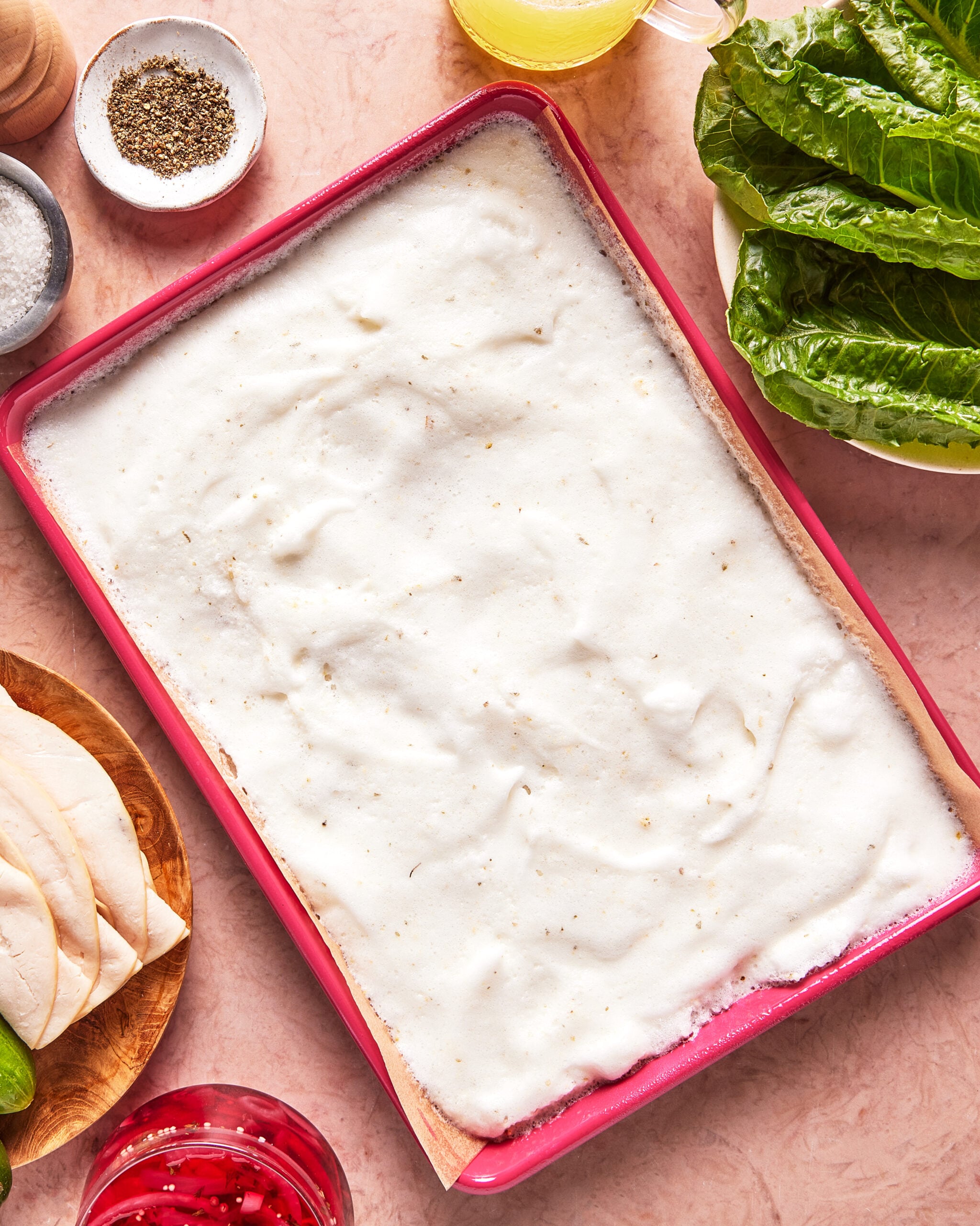 A baking dish filled with an unbaked creamy white layer sits on a table, surrounded by lettuce, sliced chicken, seasonings, and other ingredients.