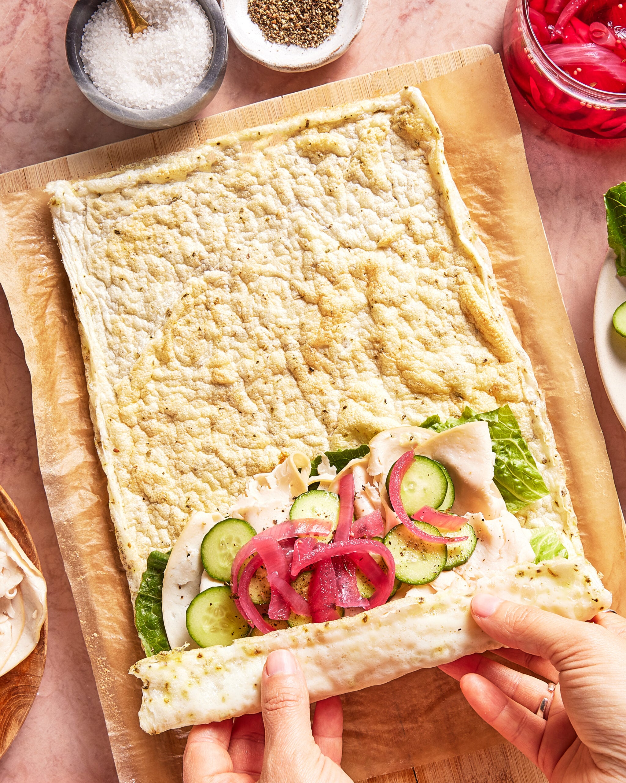 Hands rolling a flatbread wrap filled with lettuce, sliced turkey, cucumbers, and pickled red onions on parchment paper, with bowls of salt, pepper, and pickled onions nearby.