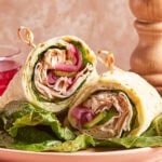 Two turkey and vegetable wraps are cut in half and stacked on a plate, secured with skewers. Visible ingredients include turkey slices, lettuce, cucumber, and pickled red onions. A pepper grinder is in the background.