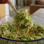 A hand holds chopsticks lifting a portion of shredded green salad with seeds from a glass bowl, in a modern, softly-lit room.