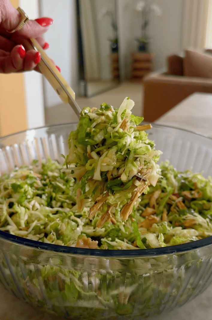 A hand holds chopsticks lifting a portion of shredded green salad with seeds from a glass bowl, in a modern, softly-lit room.