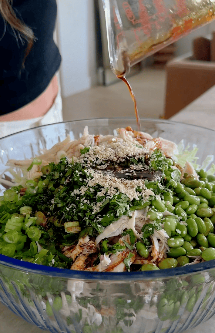A glass bowl filled with shredded chicken, green onions, edamame, herbs, and sesame seeds, with soy sauce being poured on top.