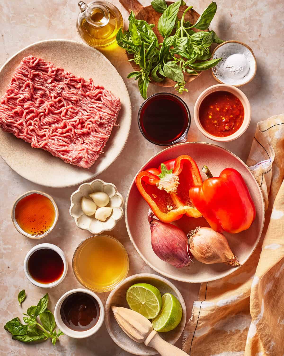 Raw ground beef, red bell peppers, shallots, garlic, limes, fresh basil, small bowls of sauces and condiments, olive oil, and salt are arranged on a light countertop with a beige cloth.