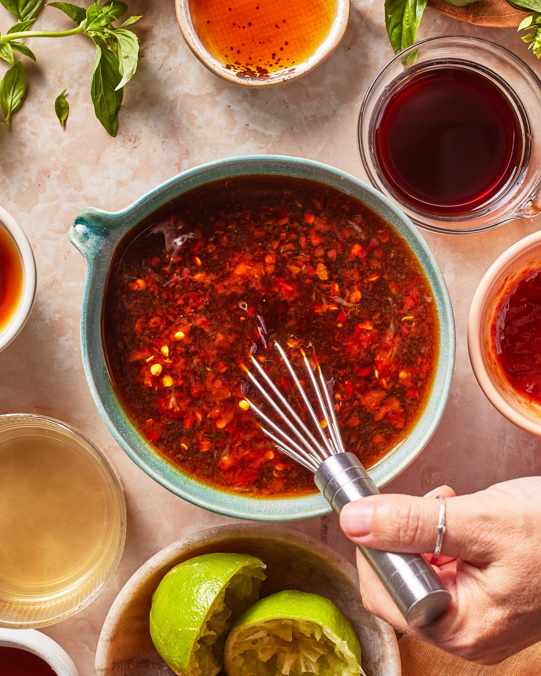 A hand whisks a spicy chili sauce in a bowl, surrounded by cut limes, herbs, and small bowls of various sauce ingredients on a light countertop.