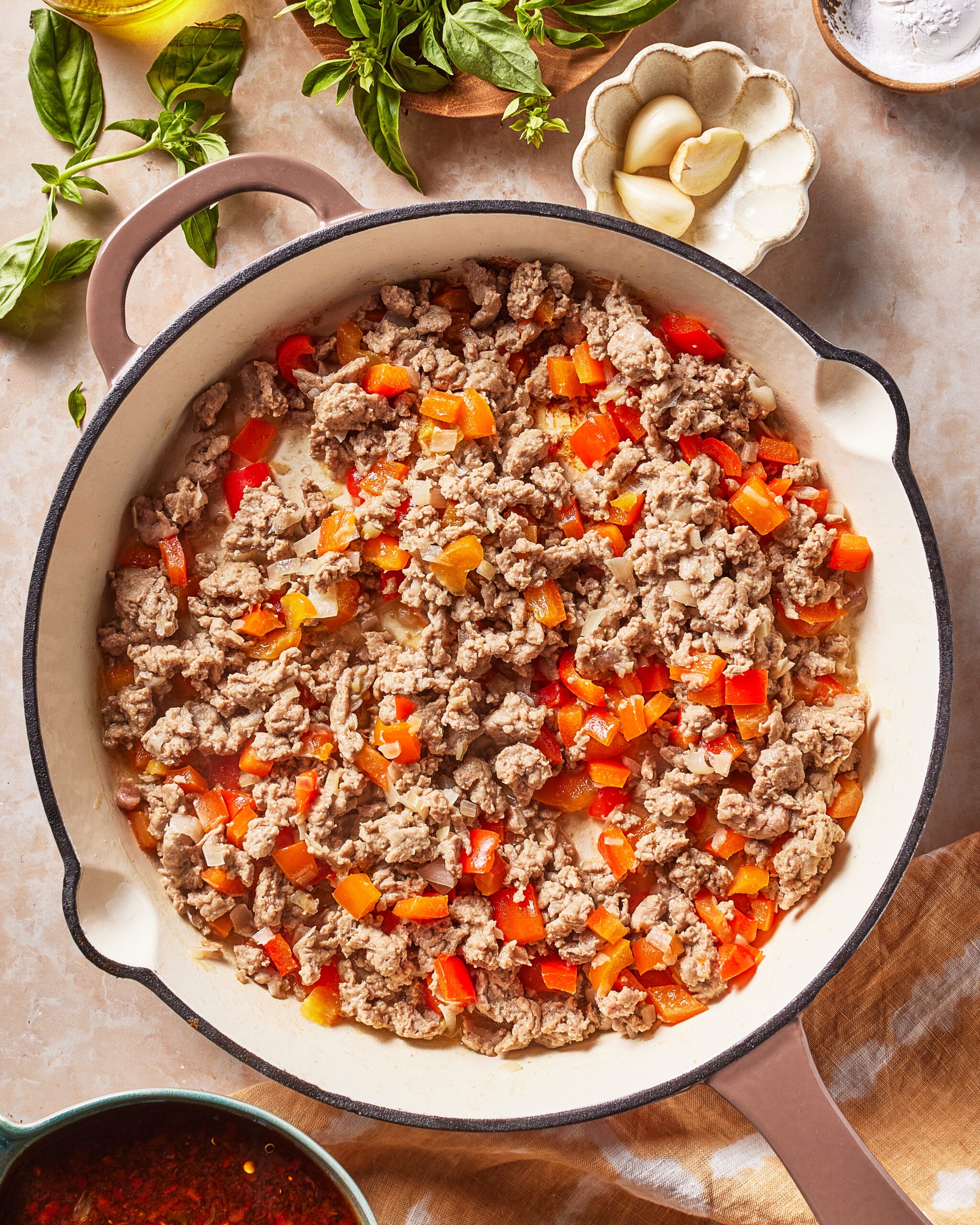 A skillet filled with cooked ground meat and diced red bell peppers, surrounded by fresh basil, garlic cloves, a bowl of sauce, and a bottle of olive oil on a marble countertop.