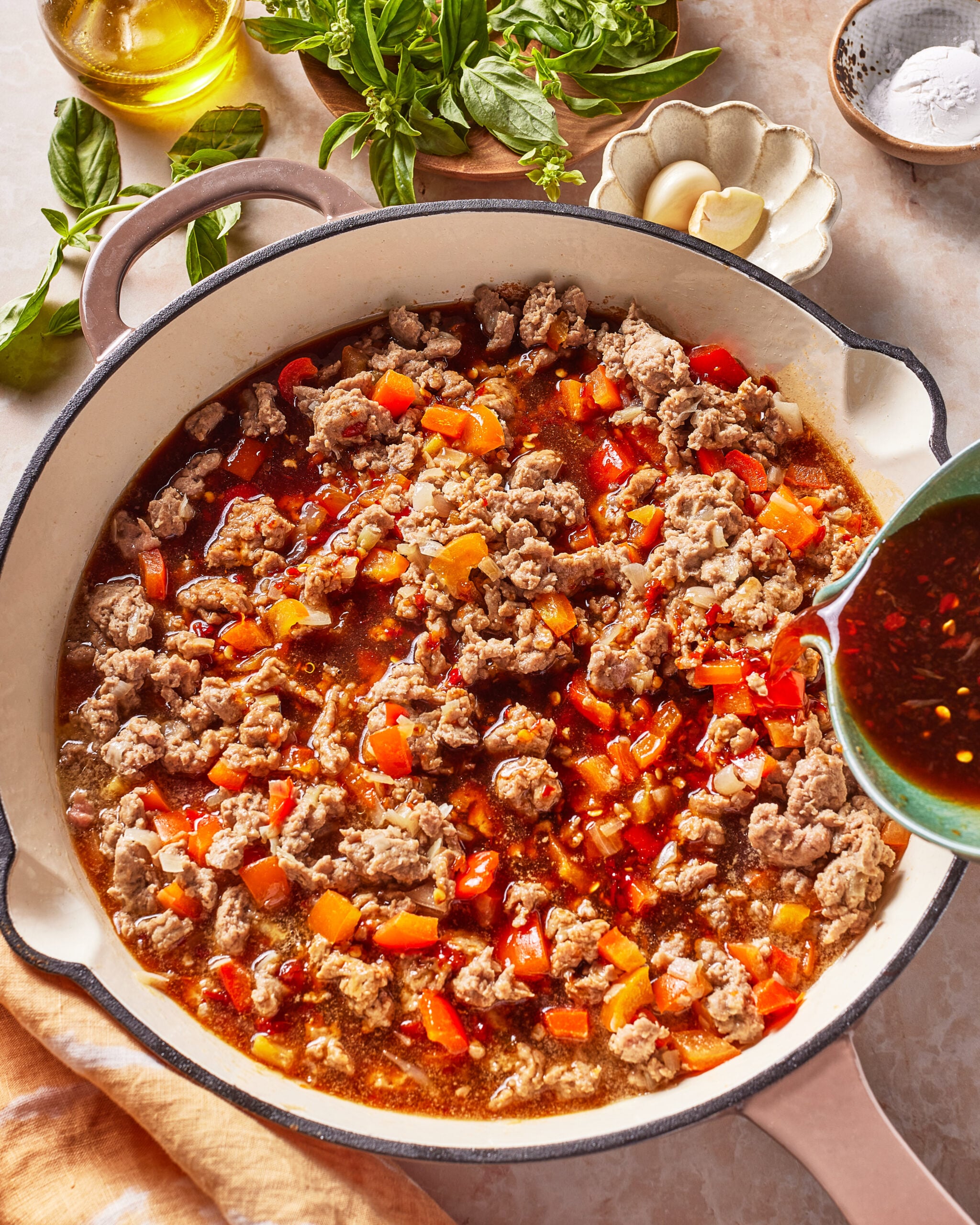 A skillet filled with cooked ground meat, diced red bell peppers, and sauce, with extra sauce being poured in. Fresh basil, garlic, oil, and seasonings are arranged nearby on the countertop.