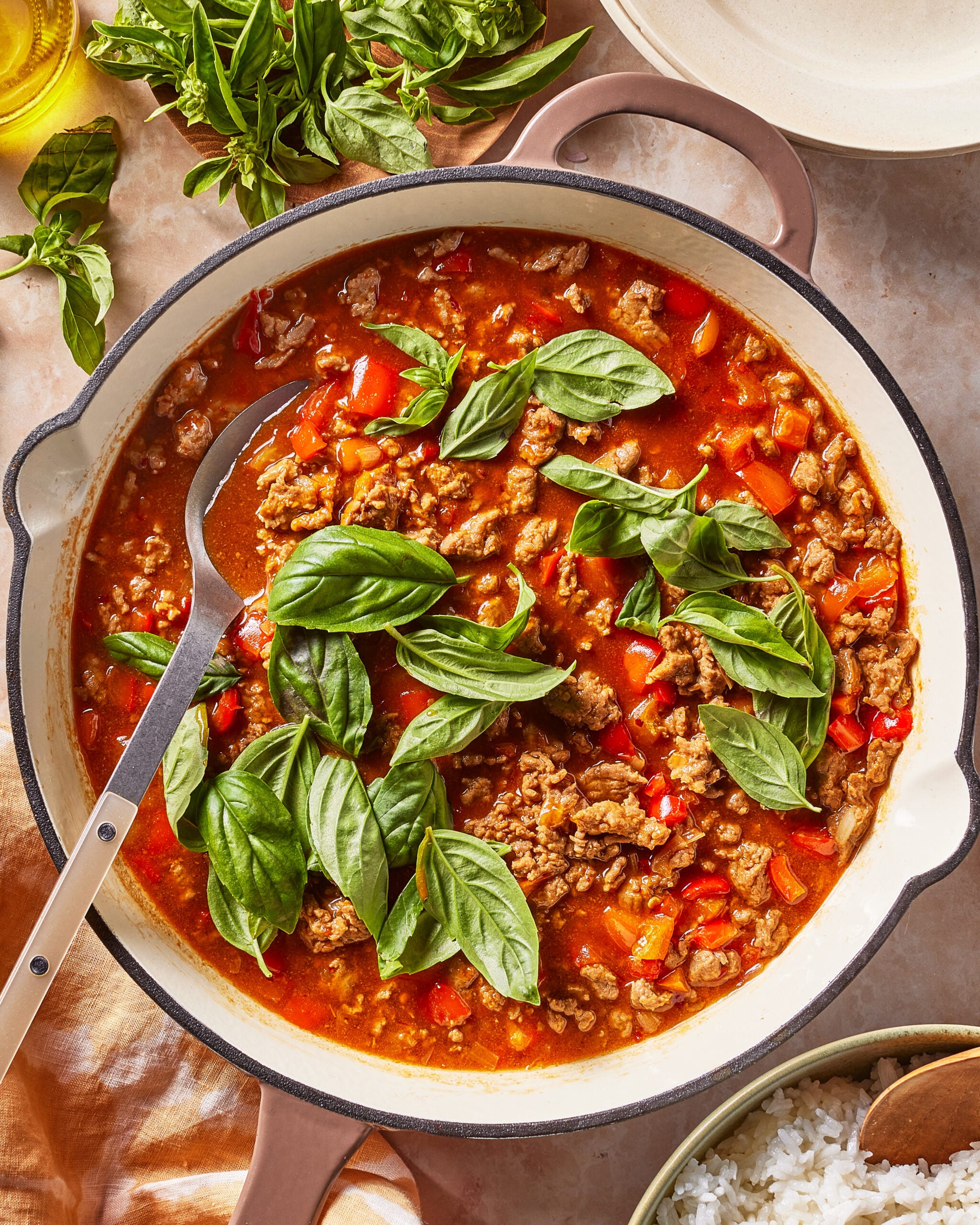 A pot of ground meat and tomato sauce with red peppers, topped with fresh basil leaves. A spoon rests inside the pot. Fresh basil, a bowl of rice, and a plate are nearby.