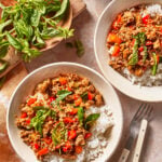 Two bowls of rice topped with stir-fried ground meat, red bell peppers, chilies, basil leaves, and sauce. Fresh basil sits nearby on a wooden plate. Fork and knife lie beside the bowls on the table.