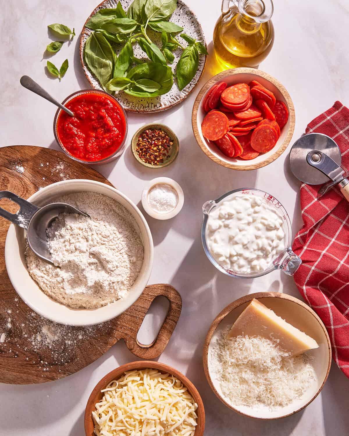 Pizza ingredients on a countertop, including flour, shredded cheese, grated parmesan, cottage cheese, pepperoni, basil leaves, tomato sauce, olive oil, and red pepper flakes, arranged in bowls and plates.