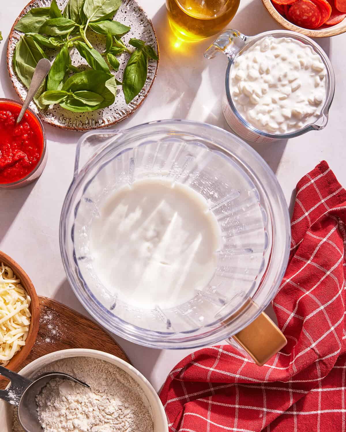 A blender of milk surrounded by cottage cheese, tomato sauce, shredded cheese, flour, fresh basil, olive oil, and a red checkered towel on a kitchen counter.