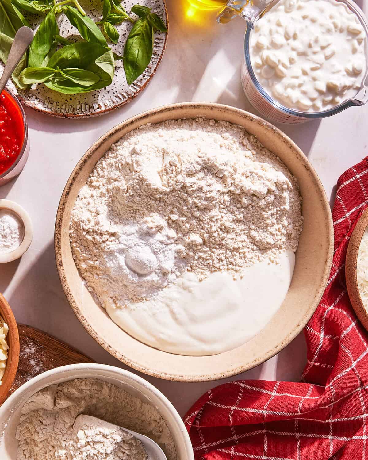A bowl of flour and yogurt sits on a table surrounded by bowls of tomato sauce, cottage cheese, fresh basil, shredded cheese, and a red kitchen towel.
