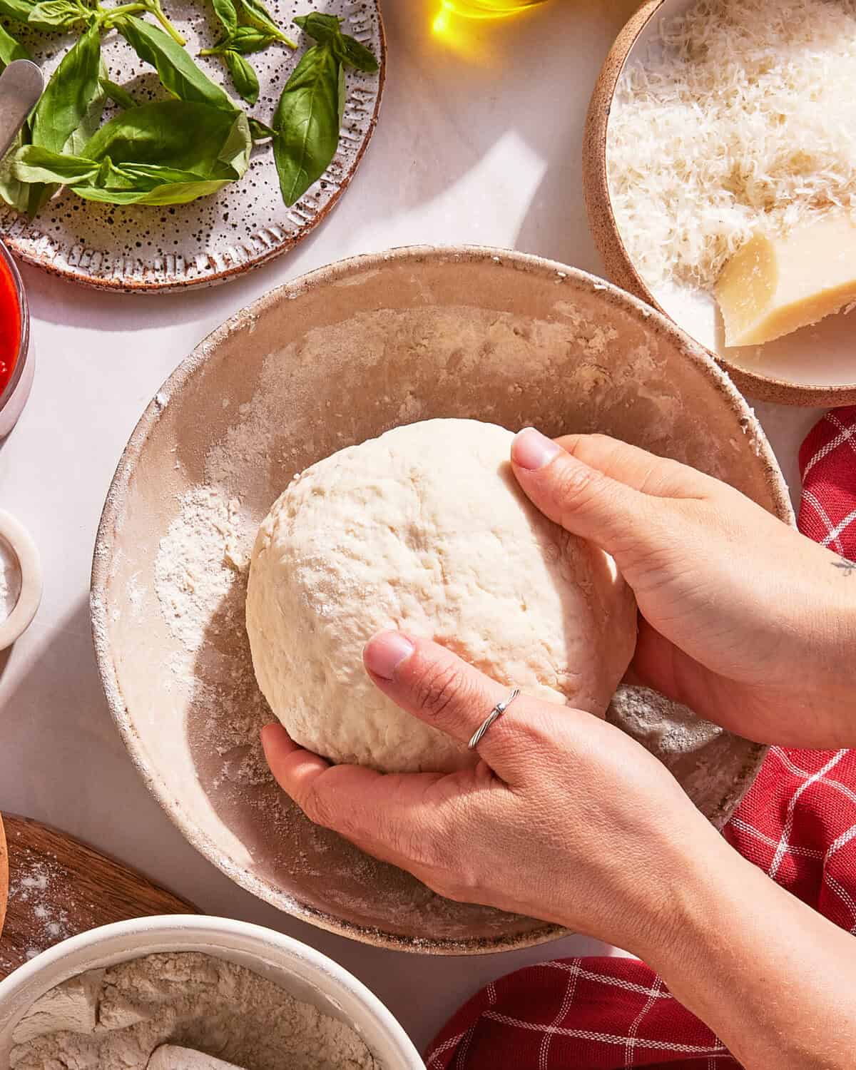 Hands knead a ball of pizza dough in a floured bowl, surrounded by plates of basil, grated cheese, a wedge of parmesan, tomato sauce, and a red checkered cloth.