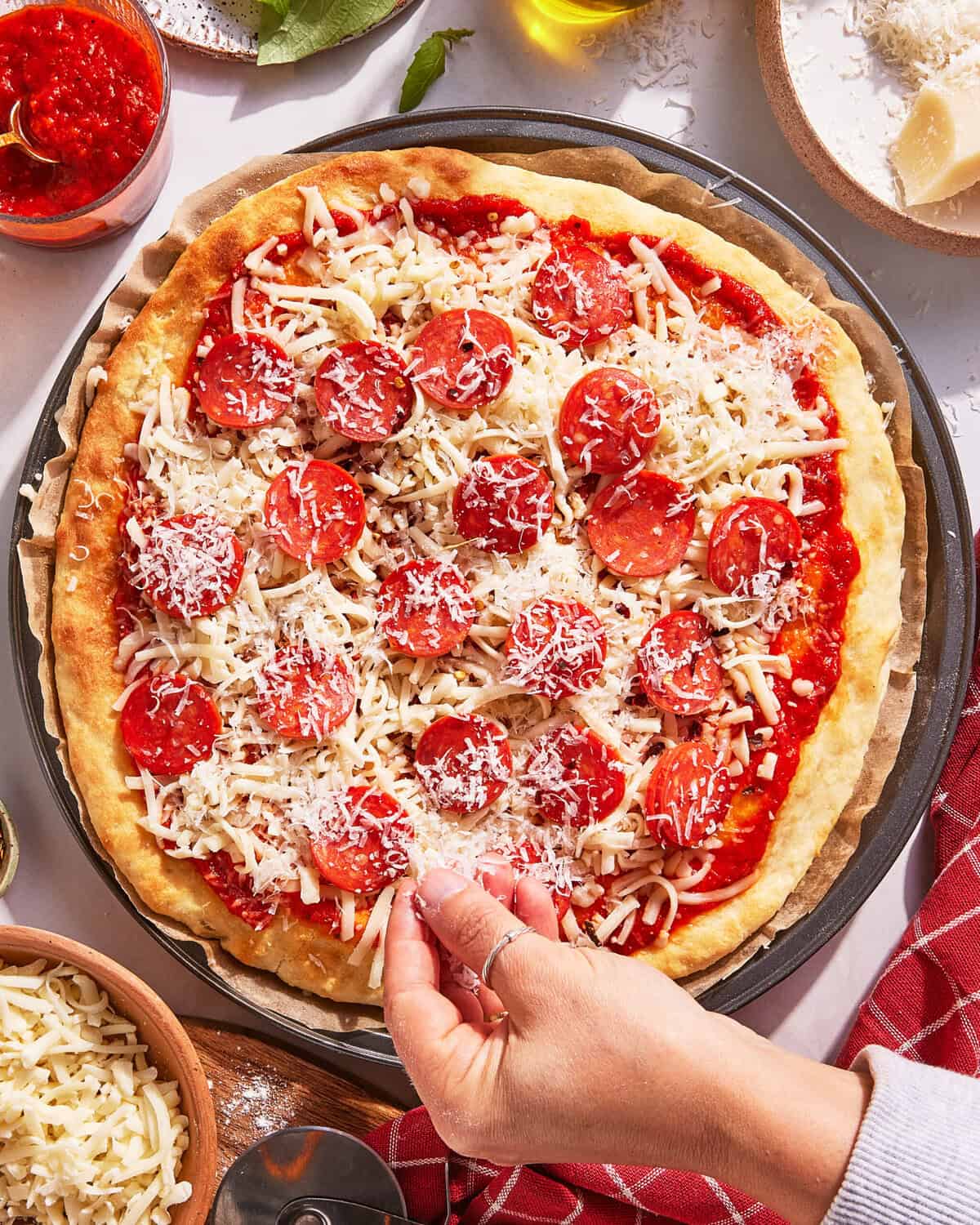 A hand sprinkles grated cheese onto a homemade pepperoni pizza with tomato sauce and shredded cheese, surrounded by bowls of cheese, sauce, and kitchen utensils.