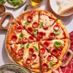 A hand takes a slice of pepperoni pizza topped with basil leaves. The pizza sits on a wooden board surrounded by cheese, olive oil, basil, red pepper flakes, and a checkered napkin.