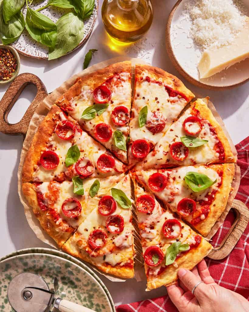 A hand takes a slice of pepperoni pizza topped with basil leaves. The pizza sits on a wooden board surrounded by cheese, olive oil, basil, red pepper flakes, and a checkered napkin.