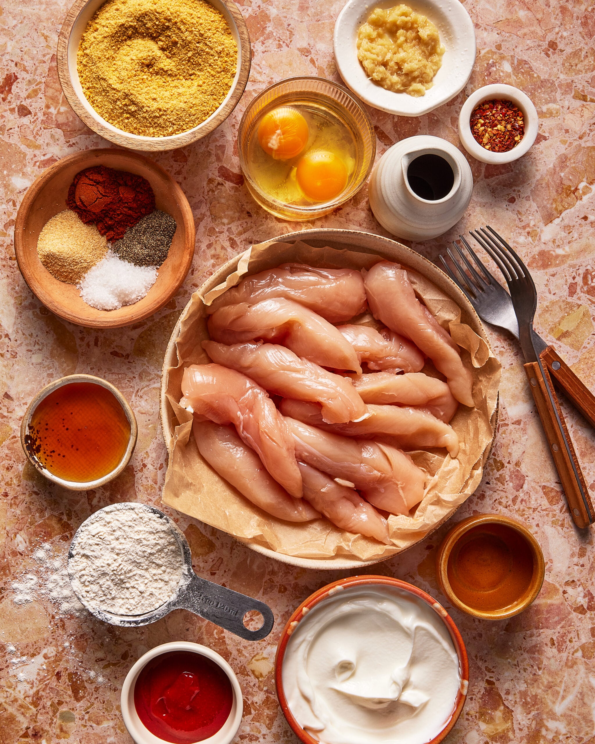 Raw chicken tenders on parchment surrounded by bowls of eggs, flour, spices, minced garlic, yogurt, honey, mustard, ketchup, and oil on a marble surface, with a fork and knife nearby.