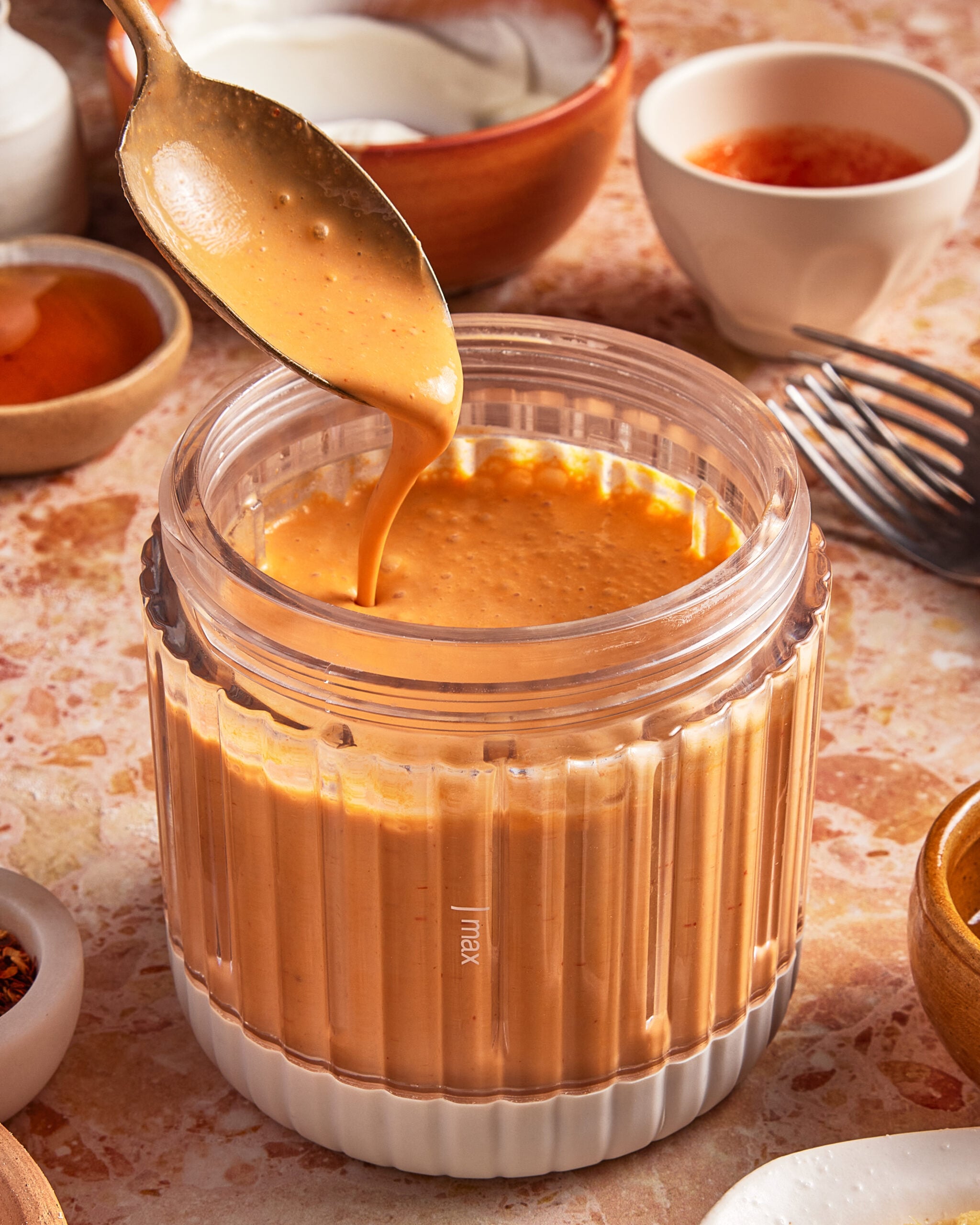A spoon drizzles creamy orange sauce into a glass blender jar, surrounded by small bowls and utensils on a textured countertop.