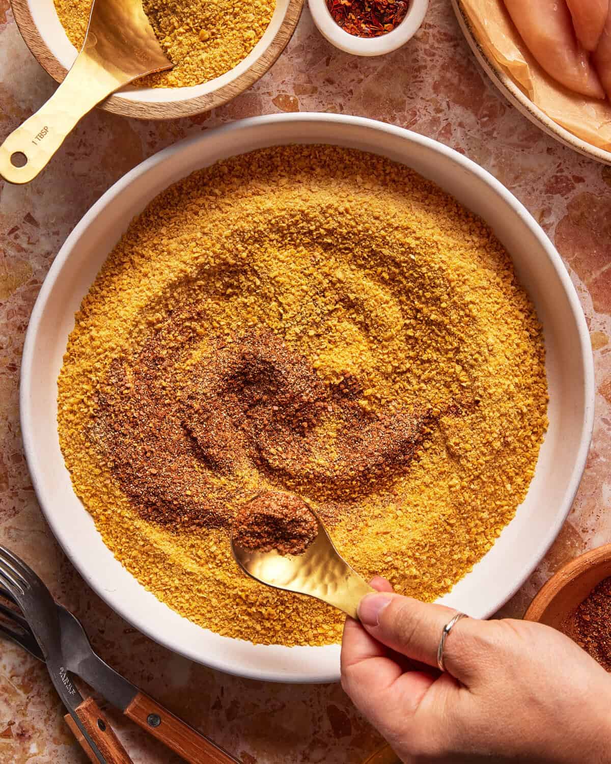 A hand stirs brown spices into a bowl of yellow breadcrumbs with a gold spoon. Other bowls and utensils are visible around on a marbled surface.