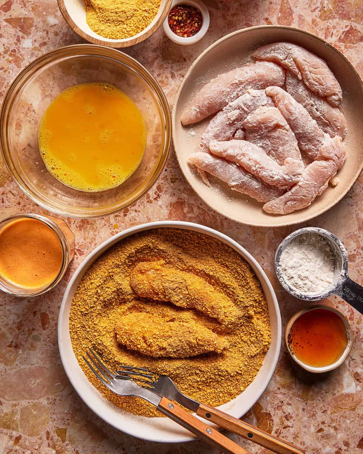 Bowls of raw chicken tenders, beaten eggs, flour, spices, and bread crumbs arranged on a countertop, with some tenders being coated in bread crumbs using a fork and knife.