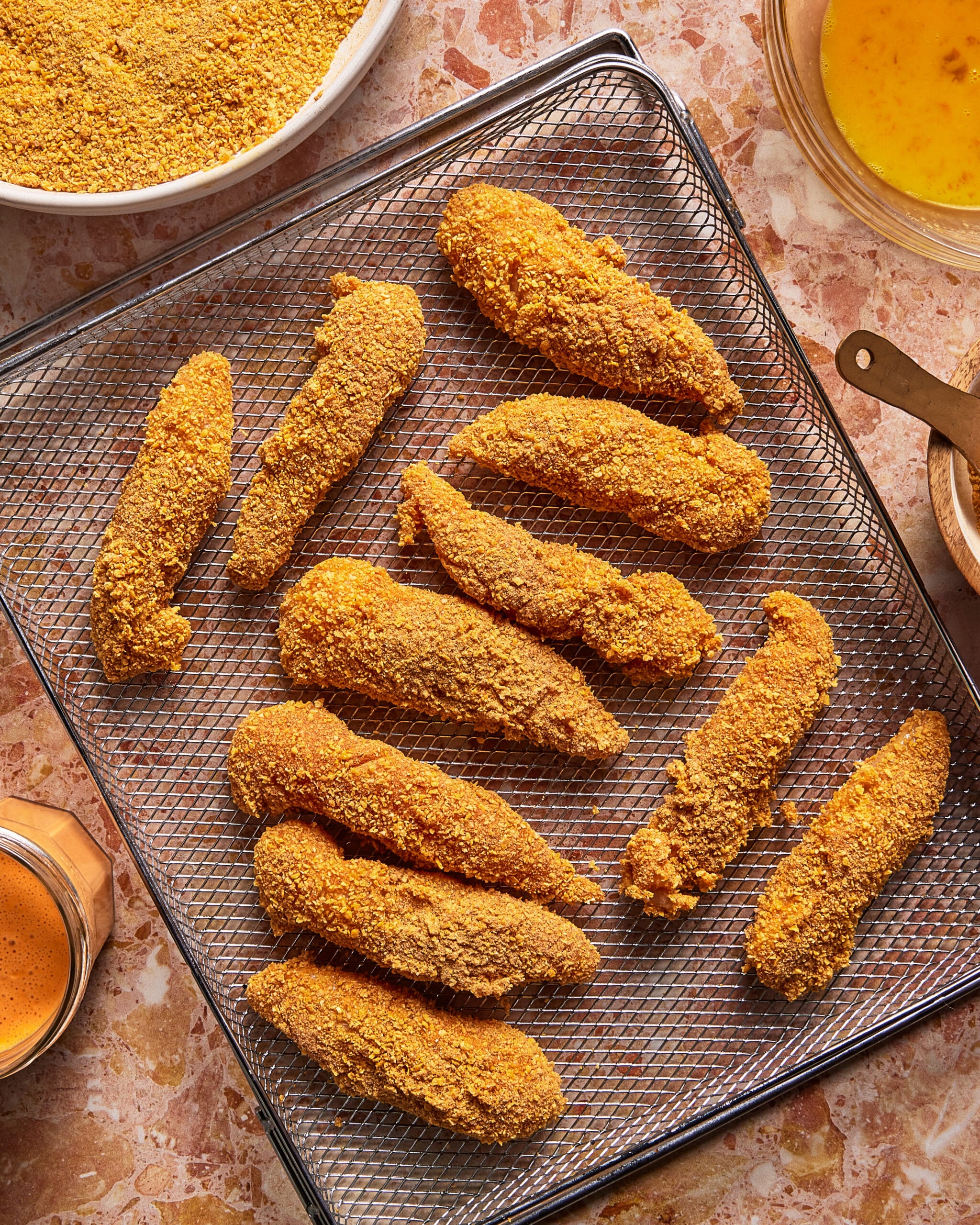 Battered, uncooked chicken tenders arranged on a wire rack, with bowls containing breading and egg wash nearby on a marbled countertop.